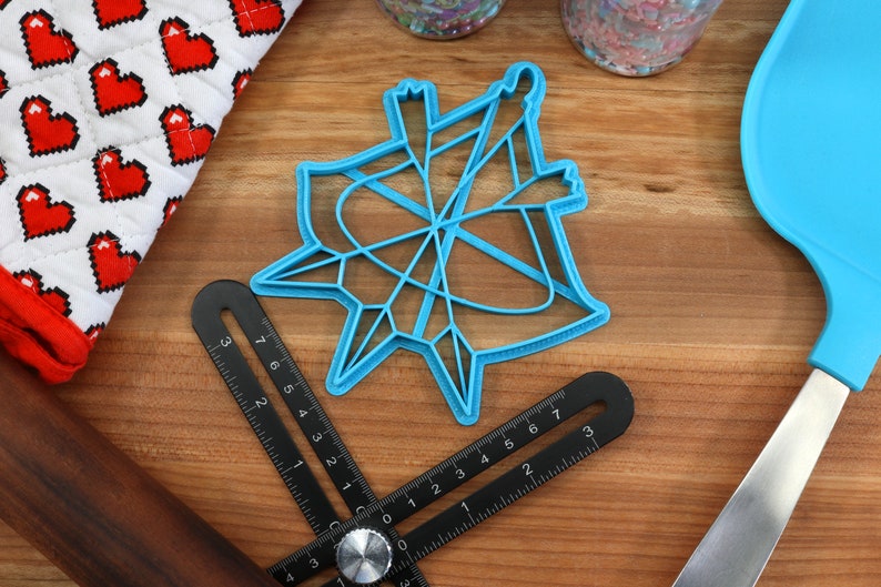 May include: A blue plastic cookie cutter shaped like a star with a geometric design. The cutter is on a wooden surface with a ruler, a rolling pin, and a spatula.