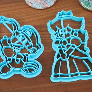 May include: Two blue plastic cookie cutters, one shaped like a princess and the other shaped like a character wearing a hat.