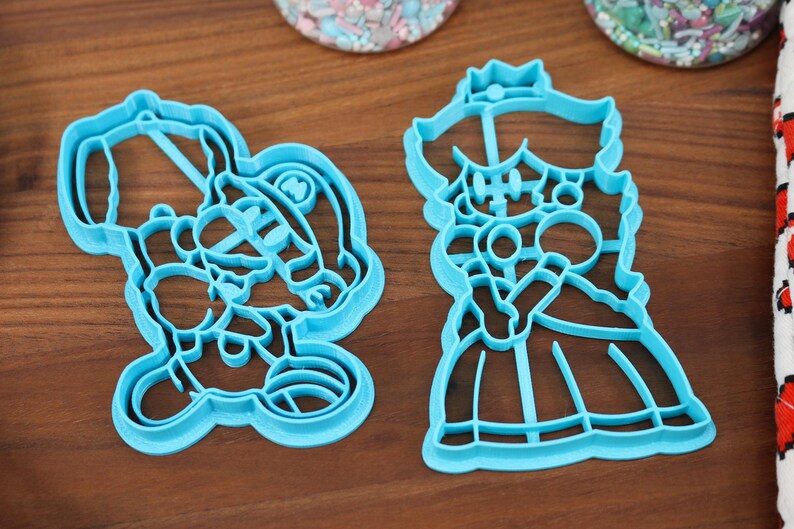 May include: Two blue plastic cookie cutters, one shaped like a princess and the other shaped like a character wearing a hat.
