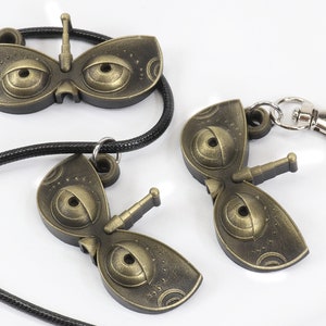 May include: Two gold-colored metal keychains shaped like a butterfly with large eyes. One keychain is attached to a black cord, while the other has a silver metal clasp.