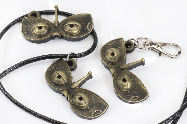 May include: Two gold-colored metal keychains shaped like a butterfly with large eyes. One keychain is attached to a black cord, while the other has a silver metal clasp.