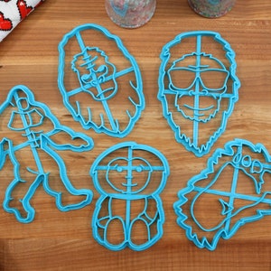 May include: Five blue plastic cookie cutters shaped like different characters. The cutters are on a wooden surface. The characters include a yeti, a sasquatch, a yeti with sunglasses, a yeti with a beard, and a yeti with a beard and sunglasses.