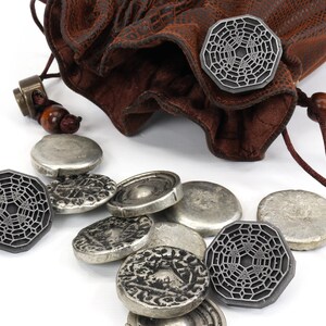 May include: A brown leather pouch with a drawstring closure, spilling out silver and gray metal tokens. The tokens feature a geometric design.