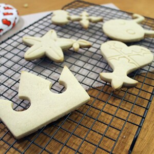 May include: Five plain sugar cookies on a black wire cooling rack. The cookies are shaped like a crown, a star, a heart, and two other shapes. The cookies are on a wooden surface.