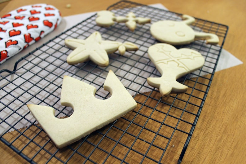 May include: Five plain sugar cookies on a black wire cooling rack. The cookies are shaped like a crown, a star, a heart, and two other shapes. The cookies are on a wooden surface.