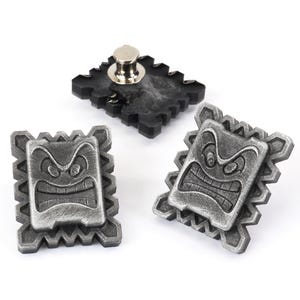 May include: Three silver-colored metal pins with a cartoon face design. The pins are shaped like a square with a jagged edge. The face on the pins has a frowning expression with a wide mouth and raised eyebrows.