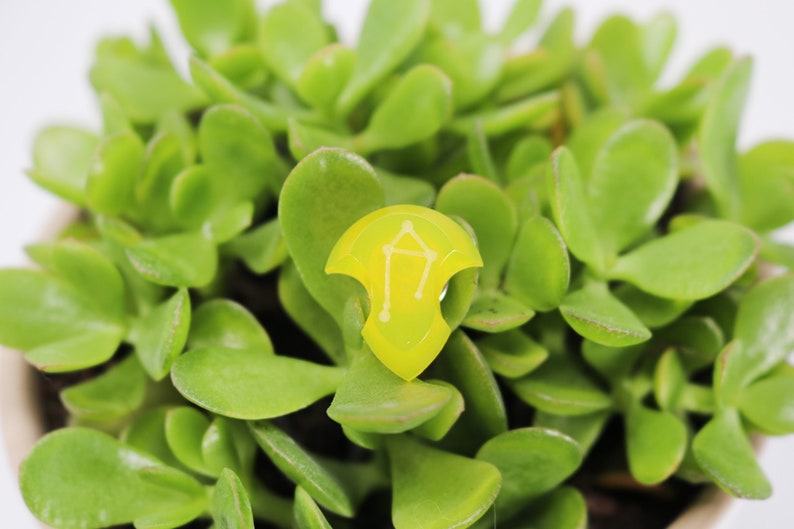 May include: A yellow resin pendant with a white outline of the Libra zodiac sign. The pendant is sitting on a bed of green succulent leaves.
