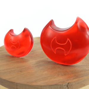 May include: Two red plastic objects shaped like a crescent moon with a stylized H design in the center. The objects are on a wooden surface.