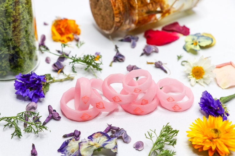 May include: A set of six pink silicone rings with a heart design. The rings are stacked on top of each other and are surrounded by dried flowers.