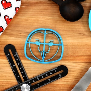 May include: A blue plastic owl cookie cutter on a wooden surface. The cutter has a detailed design of an owl's face with eyes, beak, and feathers.