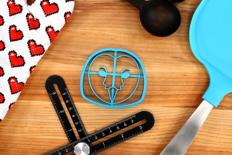 May include: A blue plastic owl cookie cutter on a wooden surface. The cutter has a detailed design of an owl's face with eyes, beak, and feathers.