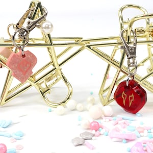 Puede incluir: Dos pinzas para el pelo doradas en forma de estrella con abalorios. Un abalorio es un coraz&oacute;n rosa con purpurina, el otro es un abalorio rojo facetado en forma de dado con un s&iacute;mbolo de flecha de doble punta.