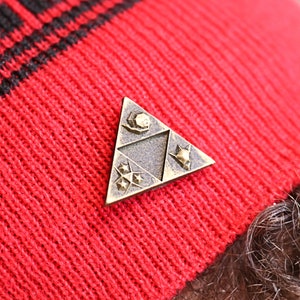May include: A gold-colored triangular pin with a stylized design of a three-pointed star, a crescent moon, and a stylized bird. The pin is attached to a red knitted hat.