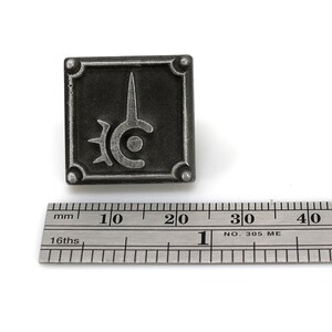 May include: A silver metal pin with a black and silver design. The design is a stylized sword with a circle and spikes around it. The pin is about 2 cm wide.