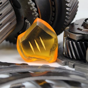 May include: An assortment of mechanical gears and components, including a translucent orange piece with three vertical slits. The gears are metallic, with various sizes and tooth designs, set against a white background.