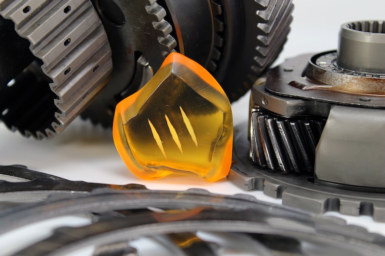 May include: An assortment of mechanical gears and components, including a translucent orange piece with three vertical slits. The gears are metallic, with various sizes and tooth designs, set against a white background.