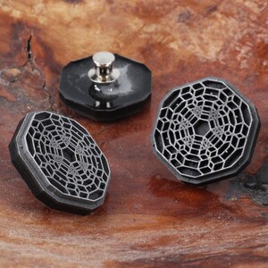 May include: Three black metal buttons with a geometric design. The buttons have a raised, textured surface and a silver metal post on the back.