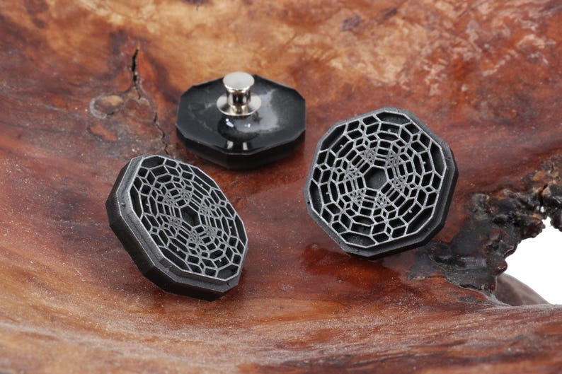 May include: Three black metal buttons with a geometric design. The buttons have a raised, textured surface and a silver metal post on the back.