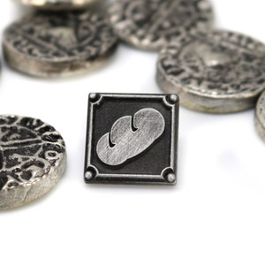 May include: A silver metal pin with a black square frame and a silver bread design inside. The pin is surrounded by several silver coins with intricate designs.