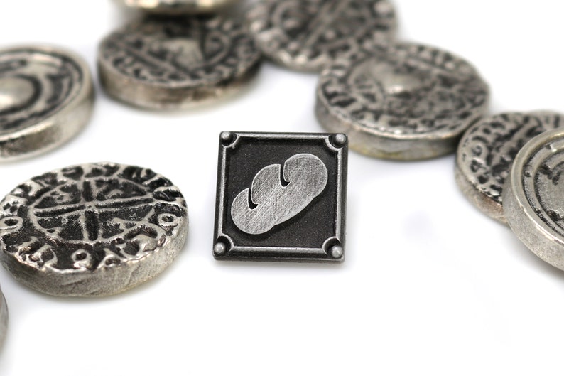 May include: A silver metal pin with a black square frame and a silver bread design inside. The pin is surrounded by several silver coins with intricate designs.