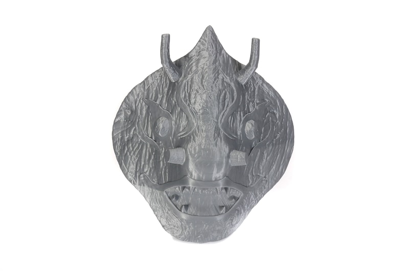Puede incluir: Una m&aacute;scara gris, texturizada, impresa en 3D con una expresi&oacute;n feroz. La m&aacute;scara tiene una parte superior puntiaguda, dos cuernos y una boca ancha y dentada.