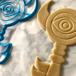 Op de afbeelding: Een blauwe plastic koekjesvormpje en een bruine suikerkoek in de vorm van een sleutel met een oogontwerp.