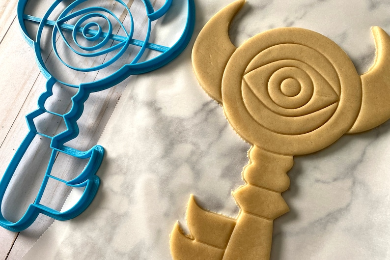 Op de afbeelding: Een blauwe plastic koekjesvormpje en een bruine suikerkoek in de vorm van een sleutel met een oogontwerp.