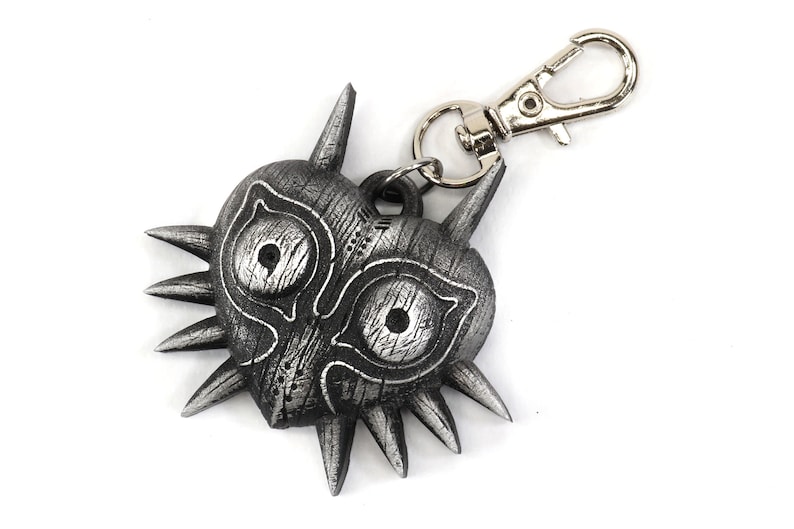 May include: A silver-colored Majora's Mask keychain with a heart-shaped design and sharp, pointed edges. The mask has two large, round eyes with a textured, wooden appearance. The keychain is attached to a silver metal ring.
