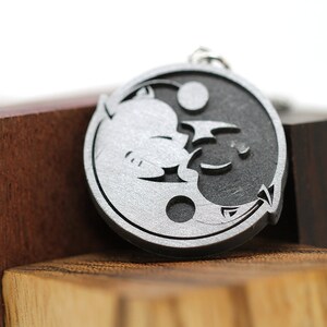 May include: A silver-colored yin yang charm with a stylized dragon and cat design. The charm is on a wooden surface.