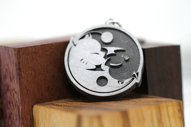 May include: A silver-colored yin yang charm with a stylized dragon and cat design. The charm is on a wooden surface.