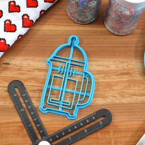 May include: A blue plastic cookie cutter shaped like a French press coffee maker. The cutter is on a wooden surface with other baking supplies.