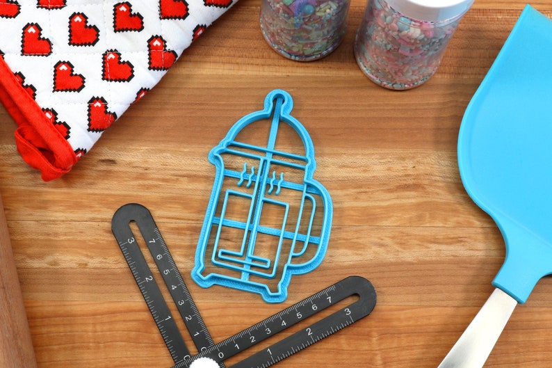 May include: A blue plastic cookie cutter shaped like a French press coffee maker. The cutter is on a wooden surface with other baking supplies.
