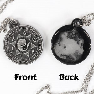 May include: A silver-toned pendant necklace with a round, double-sided charm. The front features an engraved design with text and a central figure. The back is black with a white, cloud-like pattern. The chain is also silver.