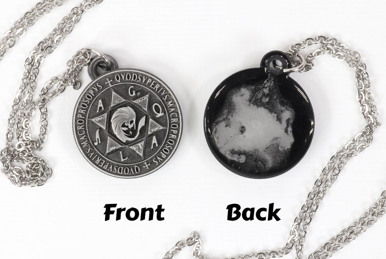 May include: A silver-toned pendant necklace with a round, double-sided charm. The front features an engraved design with text and a central figure. The back is black with a white, cloud-like pattern. The chain is also silver.
