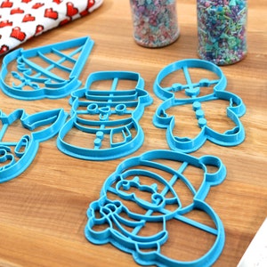 Op de afbeelding: Een set van vijf blauwe plastic kerstkoekjesvormen in de vorm van een sneeuwpop, rendier, peperkoekman, kerstman en een kerstboom. De vormen liggen op een houten oppervlak.