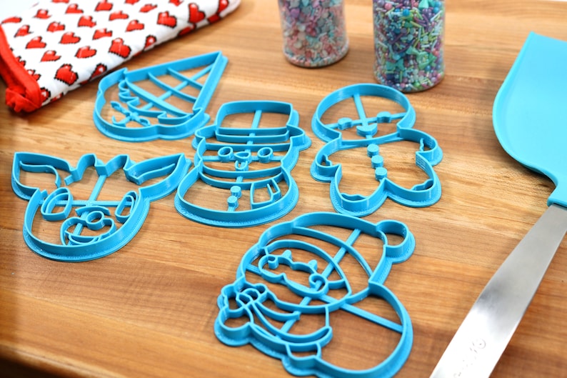 Op de afbeelding: Een set van vijf blauwe plastic kerstkoekjesvormen in de vorm van een sneeuwpop, rendier, peperkoekman, kerstman en een kerstboom. De vormen liggen op een houten oppervlak.