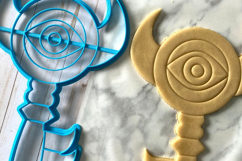 Op de afbeelding: Een blauwe plastic koekjesvormer in de vorm van een sleutel met een oogontwerp. Een koekje wordt uitgestoken met de vormer.