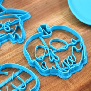May include: Three blue plastic cookie cutters shaped like a witch, a skull, and a pumpkin. The cutters are on a wooden surface.