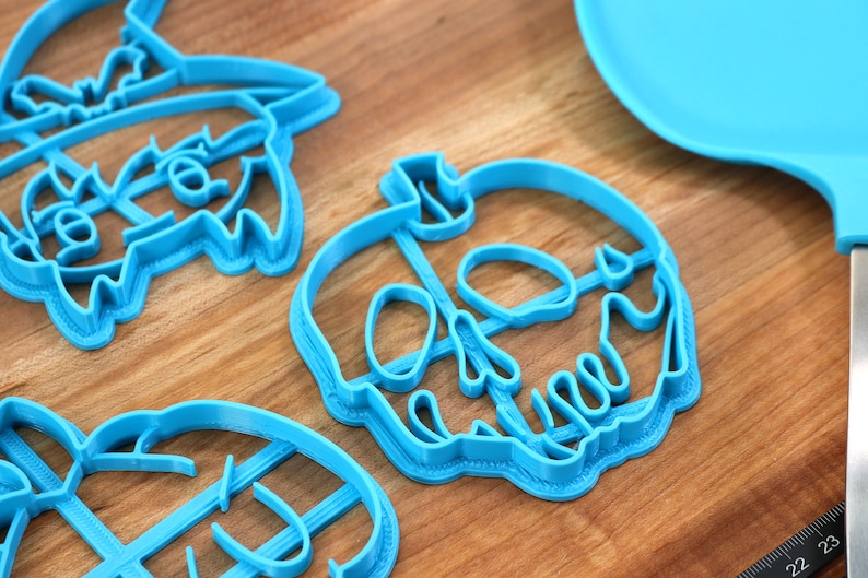 May include: Three blue plastic cookie cutters shaped like a witch, a skull, and a pumpkin. The cutters are on a wooden surface.