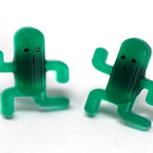 May include: Two green plastic figurines shaped like cacti with arms and legs. The cacti have simple facial features and are in a playful pose.