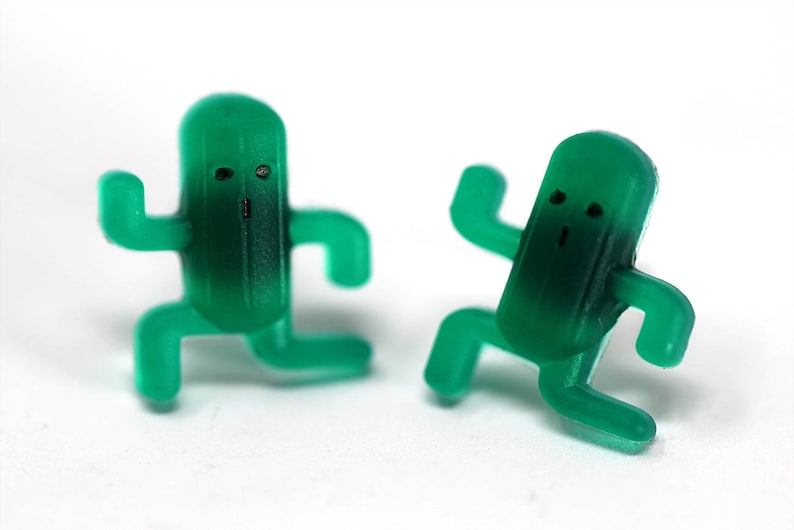 May include: Two green plastic figurines shaped like cacti with arms and legs. The cacti have simple facial features and are in a playful pose.
