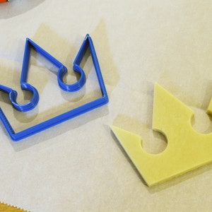 May include: A blue plastic crown cookie cutter and a yellow dough crown cutout on a white parchment paper surface.