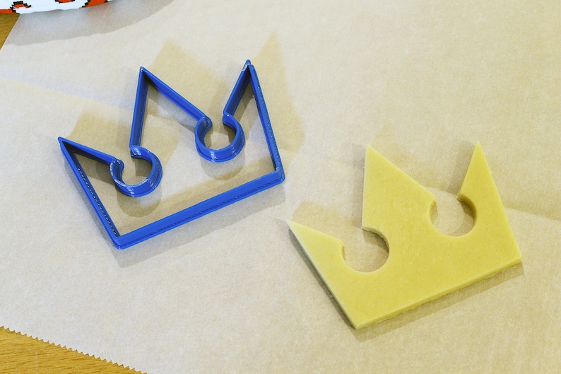 May include: A blue plastic crown cookie cutter and a yellow dough crown cutout on a white parchment paper surface.
