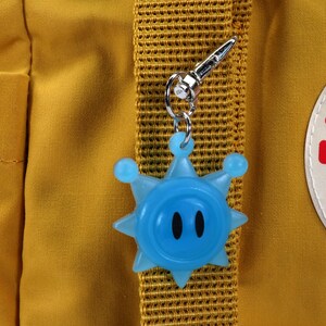 May include: A blue star-shaped keychain with a face and two black eyes attached to a yellow backpack with a Fj&auml;llr&auml;ven logo.