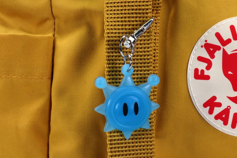 May include: A blue star-shaped keychain with a face and two black eyes attached to a yellow backpack with a Fj&auml;llr&auml;ven logo.