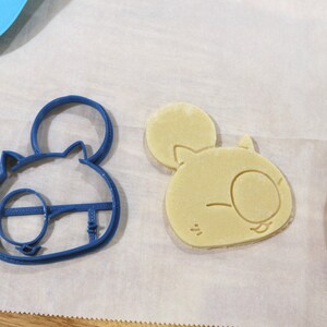 May include: A blue plastic cookie cutter shaped like a cartoon animal with large ears and a single eye. Next to it is a light brown cookie that has been cut out using the cookie cutter.