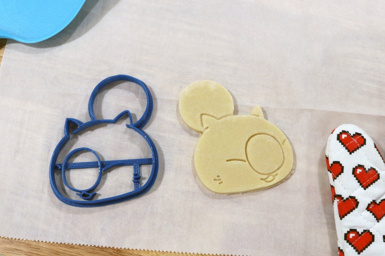 May include: A blue plastic cookie cutter shaped like a cartoon animal with large ears and a single eye. Next to it is a light brown cookie that has been cut out using the cookie cutter.