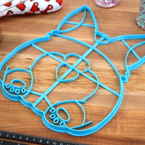 May include: A blue plastic cookie cutter shaped like a cartoon dog's face. The cutter has a heart-shaped nose and paw prints on the cheeks.