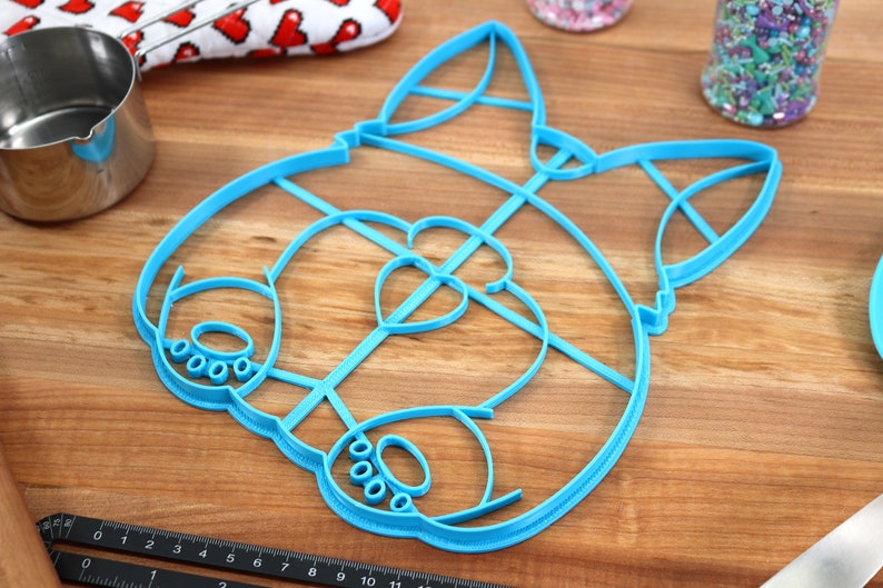 May include: A blue plastic cookie cutter shaped like a cartoon dog's face. The cutter has a heart-shaped nose and paw prints on the cheeks.