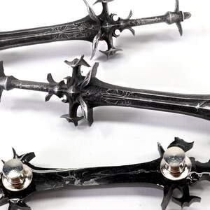 May include: Three silver-colored sword pins with intricate details and a black finish. The pins have a sharp point and a decorative design.